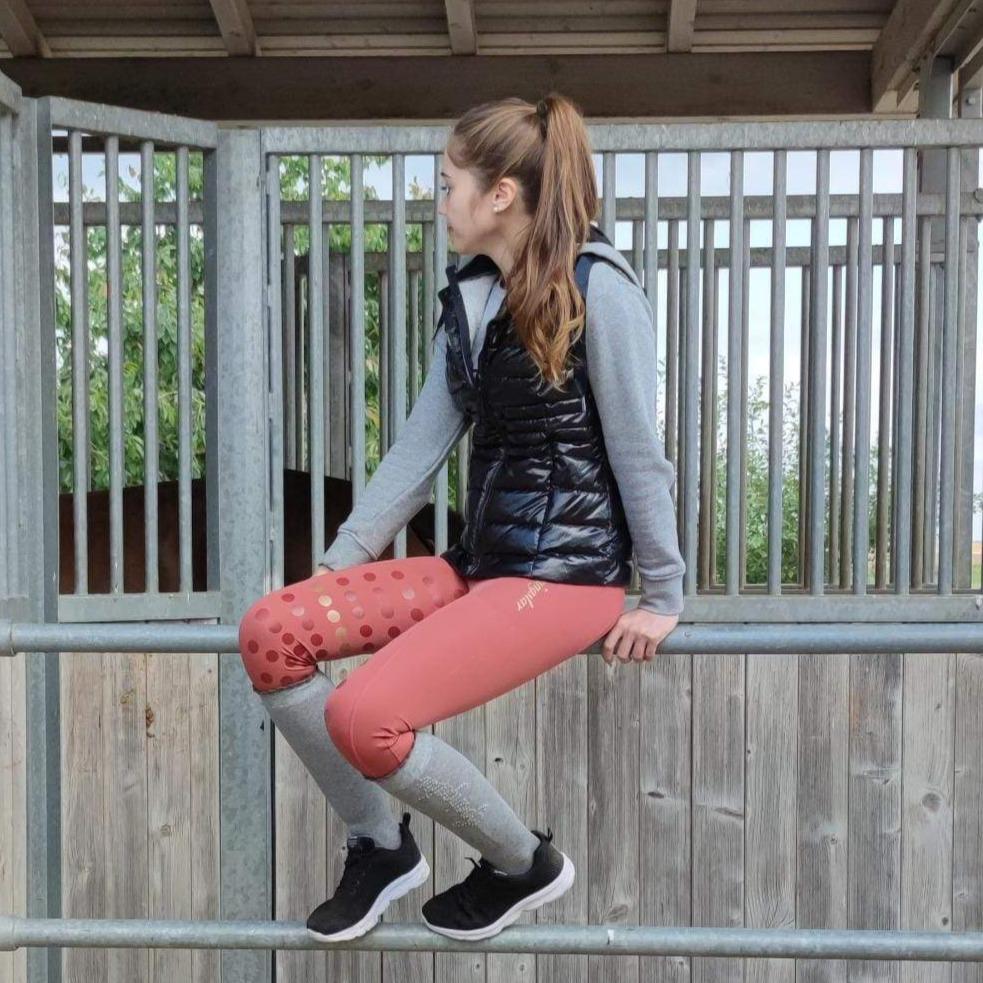 frau in pferdestall sitzt auf zaun mit schwarzer lackweste und grauem hoodie, reitleggings mit handytasche in trendfarbe marsala mit gold besatz und schwarzen sneaker schuhen mit grauen glitzer reitsocken von springstar