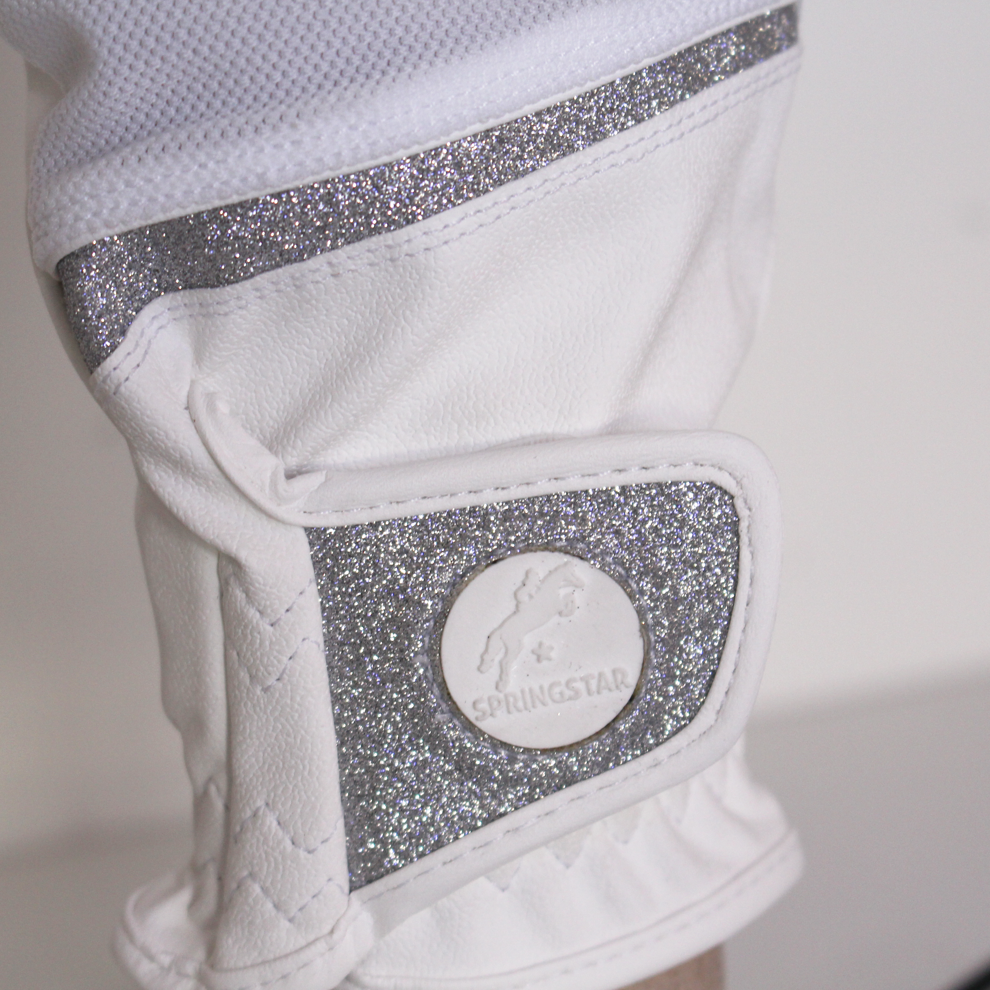 Nahaufnahme des weißen Springstar Reithandschuhs Glory 2.0 mit silbernem Glitzerband und rundem Springstar-Logo auf dem Klettverschluss.