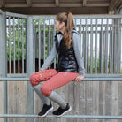 frau in pferdestall sitzt auf zaun mit schwarzer lackweste und grauem hoodie, reitleggings mit handytasche in trendfarbe marsala mit gold besatz und schwarzen sneaker schuhen mit grauen glitzer reitsocken von springstar